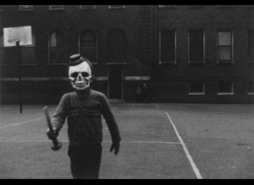 Front view of a person wearing a skeleton mask, on a school playground with a bat in their hand - black & white image