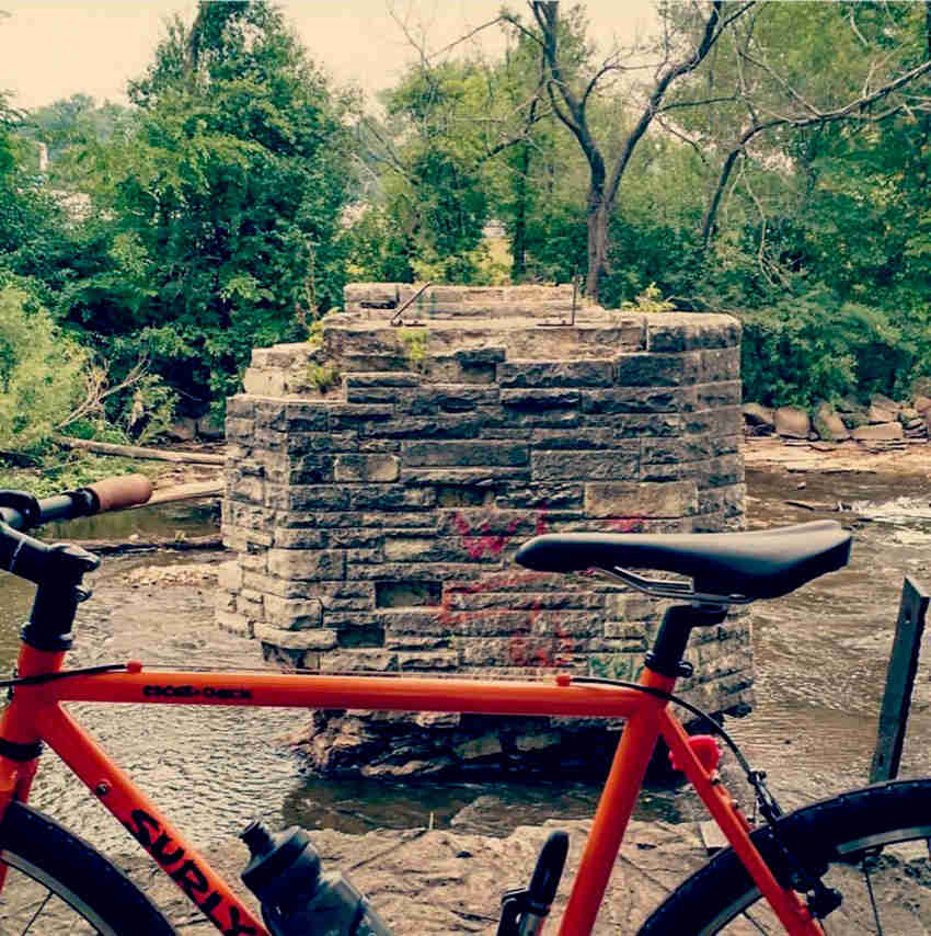 Cropped view of the upper half of an orange Surly Cross Check bike, parked in front of a stone structure in water 