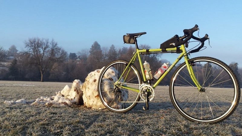 Right side view of a lime Surly Cross Check bike, in a grass field, next to a large snowball, with trees in background