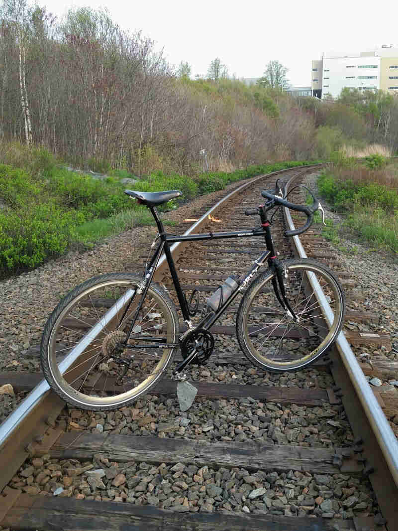 Right side view of a black Surly Cross Check bike, parked between railroad tracks that lead into the trees