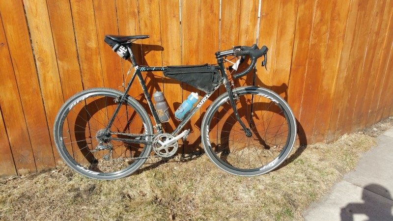 Right side view of a black Surly Cross Check bike with gear packs, parked against a wood fence wall, on grass