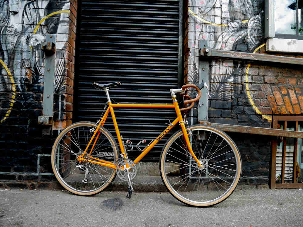 Right side view of an orange, Surly Cross Check bike, parked across a steel door on a brick building