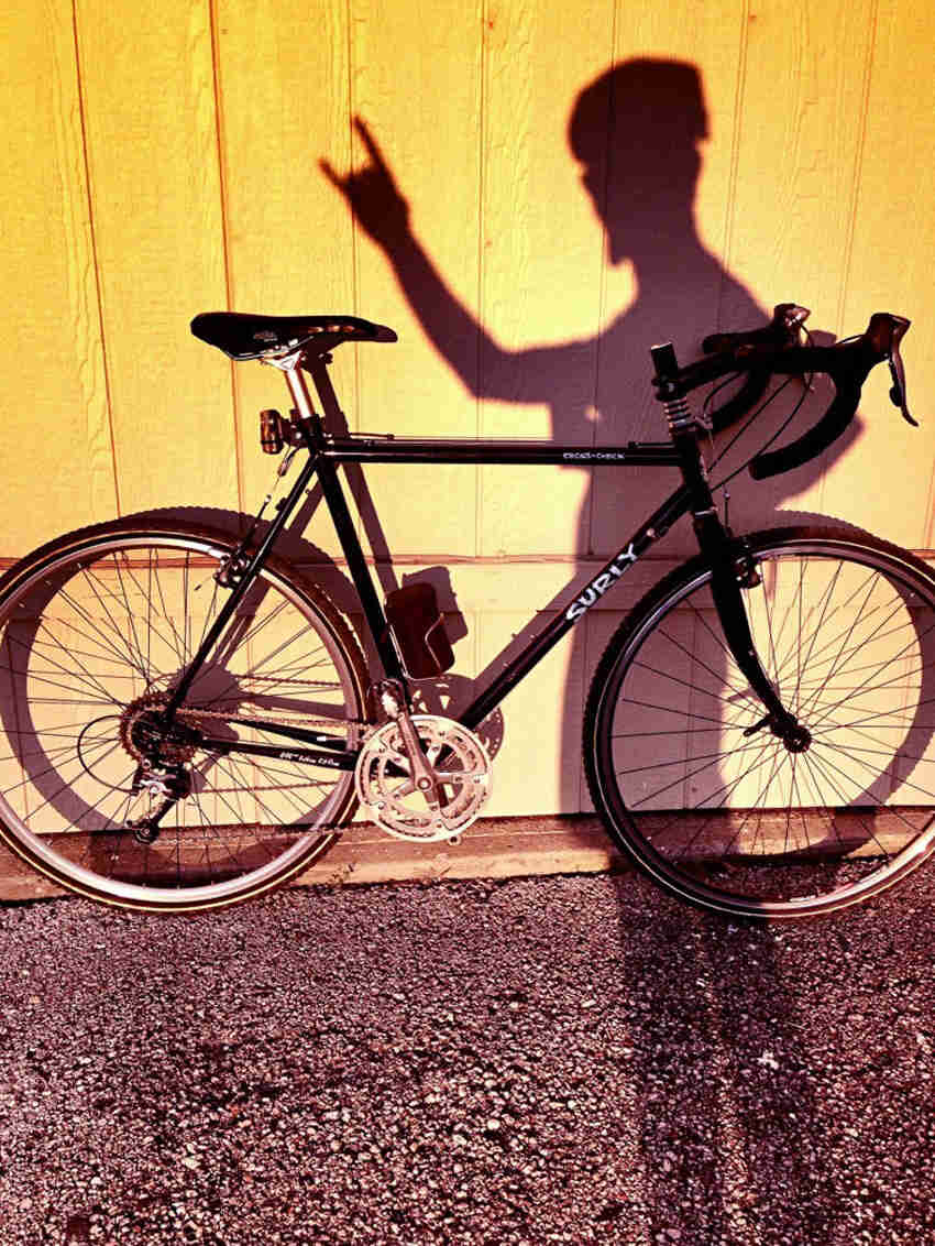 Right side view of a black Surly Cross Check bike, parked against a yellow wood wall, with a cyclist's shadow on it