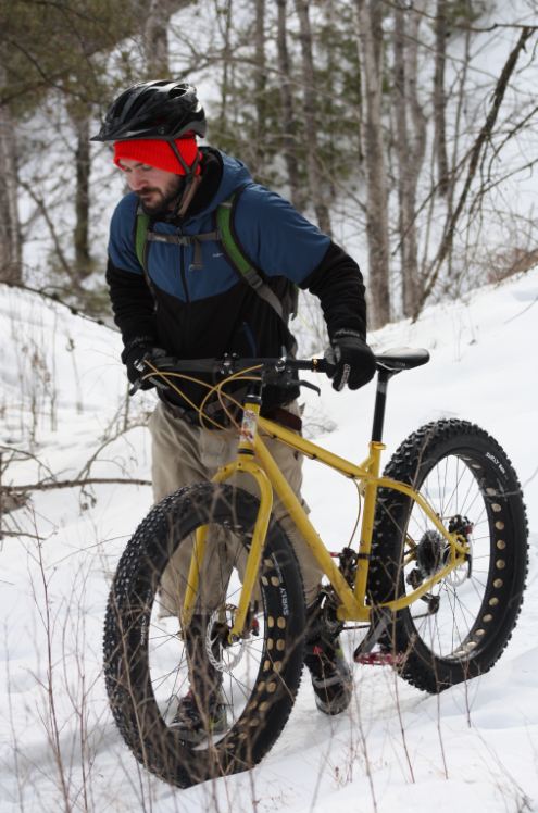 Front, left side view of a yellow Surly fat bike, with a cyclist walking it on a snow covered trail in the woods