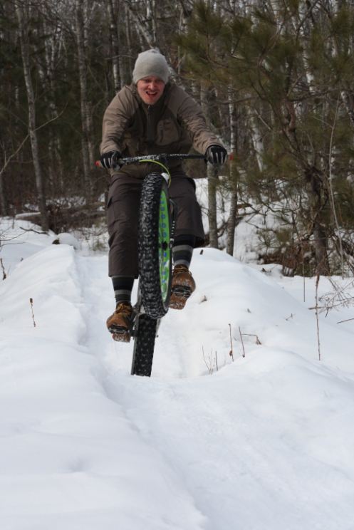 Front view of a cyclist, riding a wheelie on a white Surly Pugsley fat bike, on a snow covered trail in the woods
