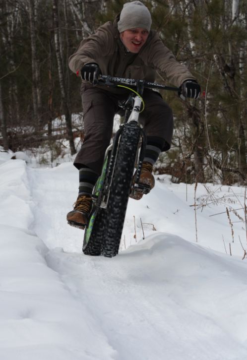 Front view of a cyclist, riding a white Surly Pugsley fat bike, down a snow covered trail in the woods