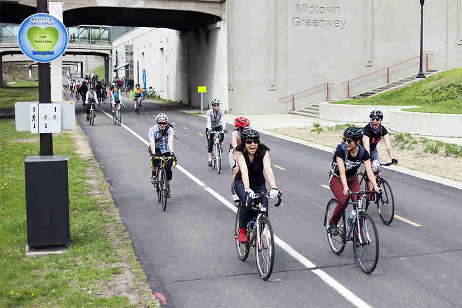 Front, right side view of a group of cyclists, riding their bikes down a paved bike street, with skywalks in background