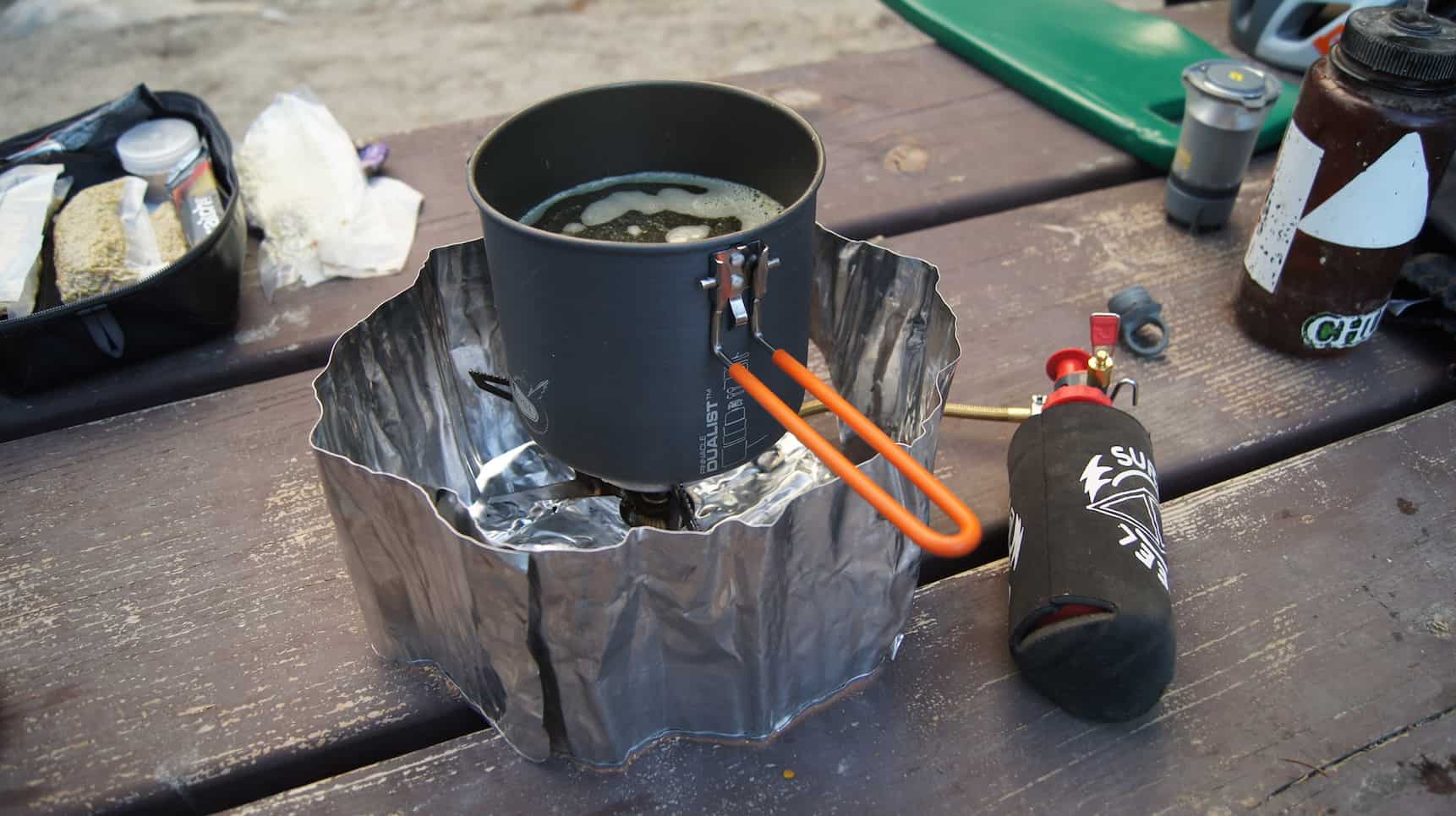 A cup of soup on top of a camp burner, with ingredients behind, on top of a wood board table