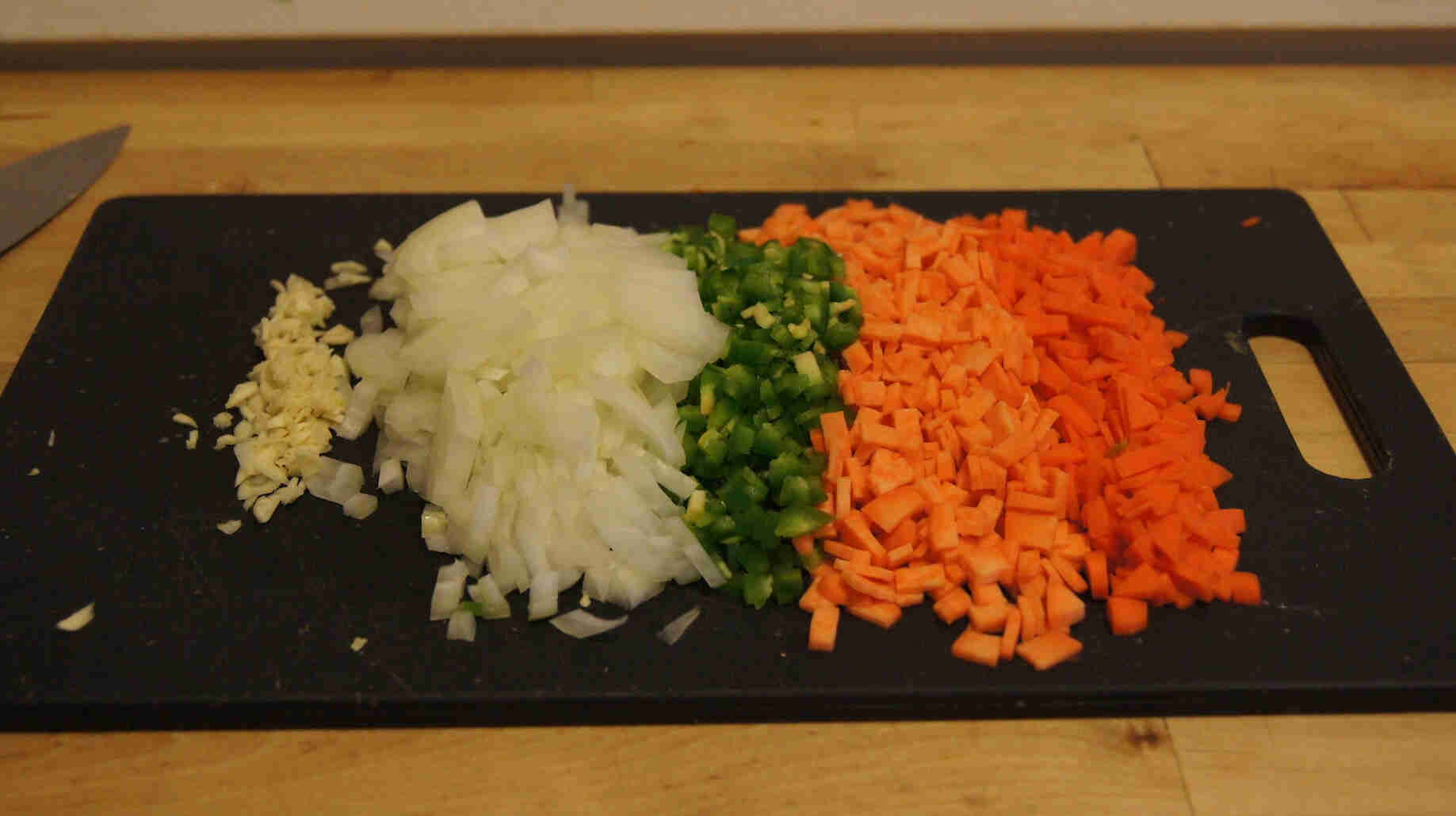 A black cutting board with diced vegetables, on top of a wood countertop