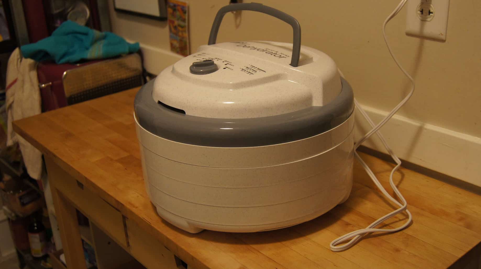 A white food dehydrator on top of wood table is plugged in to the wall outlet behind