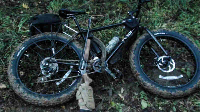 Downward, right side view of a black Surly fat bike with a rifle laying across it, lying in the weeds