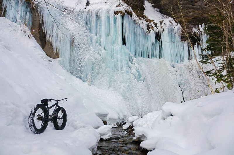 Rear right side view of a Surly fat bike in the snow, facing a frozen waterfall, on the side of a narrow stream