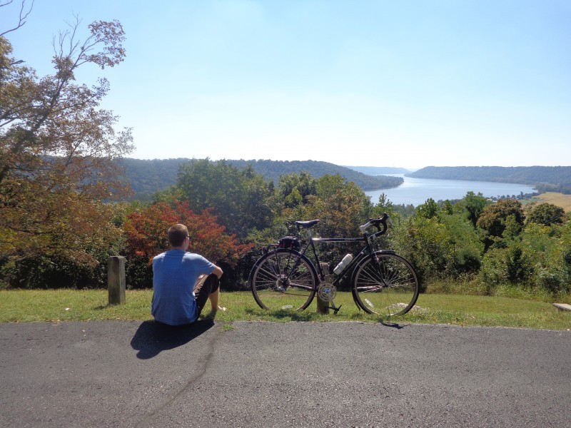 Right side view of a Surly bike, with a cyclist sitting next to it, overlooking a tree covered river valley