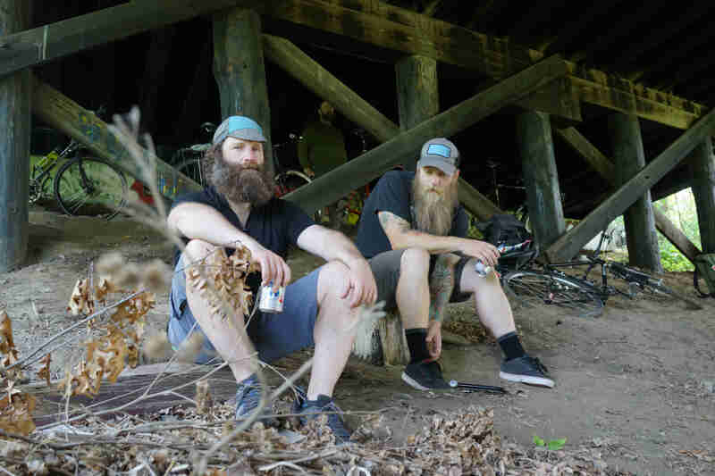 Front view of 2 bearded men, sitting on a dirt mount under a bridge, with their bikes behind them