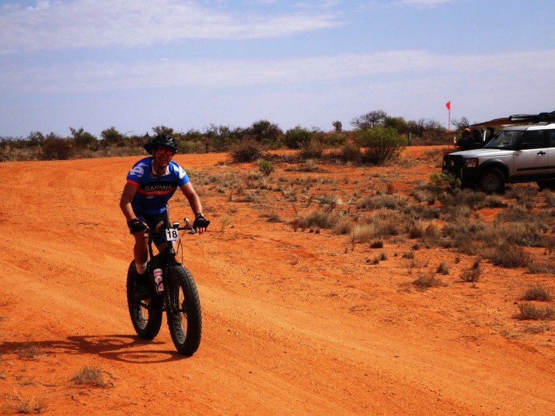 Front, right side view of a cyclist, riding a Surly fat bike with a race number in front, on a red desert road 