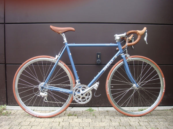 Right side view of a blue Surly Pacer bike with brown tires, seat and handlebar, parked in front of a garage door