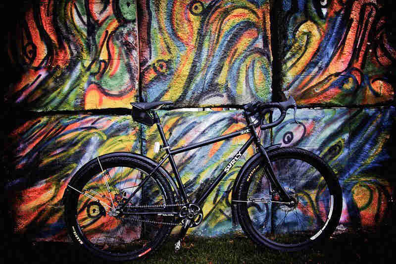 Right profile of a black Surly bike, standing in grass, against a cement block wall with a colorful mural painted on it