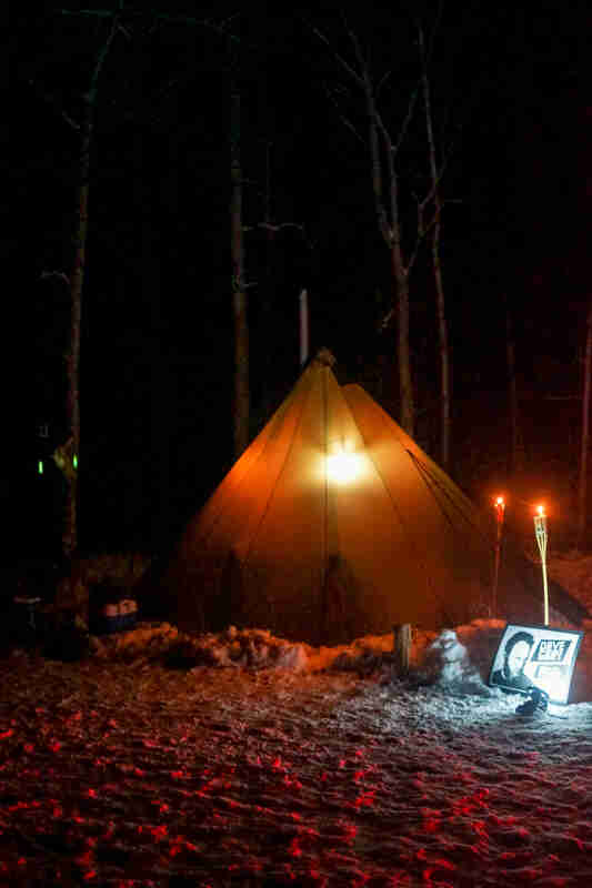 A teepee lit up with a light inside, on a snowy campsite, in the woods at night