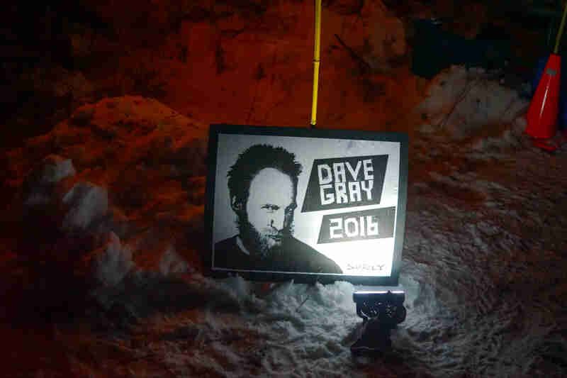 Downward view of a framed Dave Gray 2016 poster, sitting in snow with a light shining on it at night