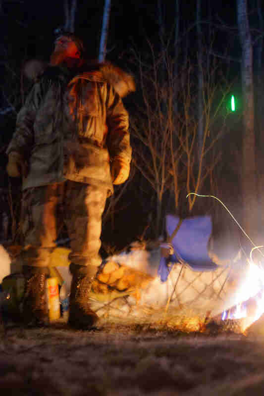 Upward view of a person, wearing winter outerwear, standing in snow next to a campfire at night