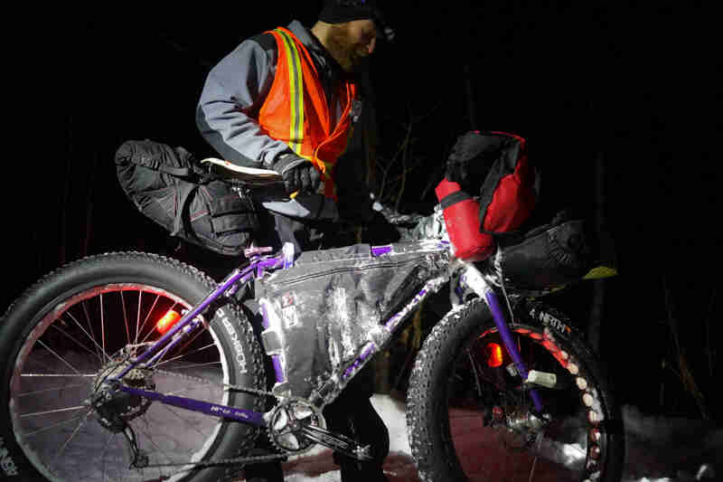 Right side view of a purple Surly Pugsley fat bike, with a cyclist standing on the left side, in the snow at night