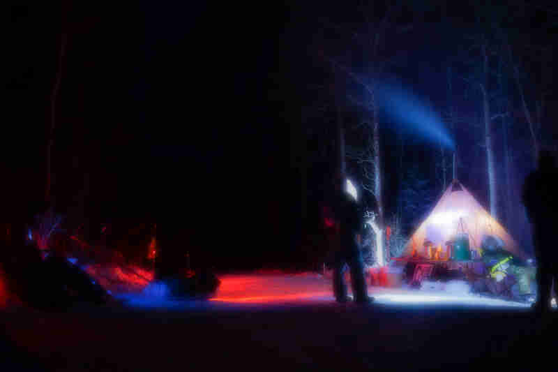 Blurry rear view of a person standing on snow, in from of a lit up teepee, in the woods at night