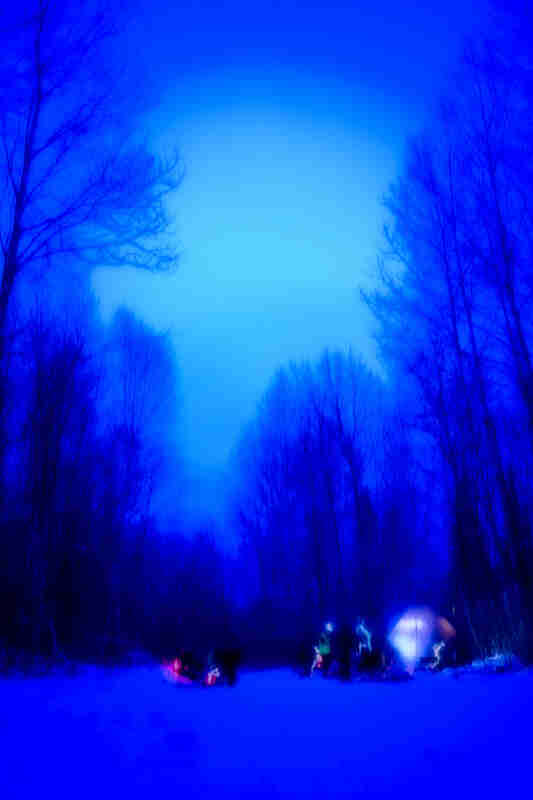 Blurry blue saturation view of a campsite in the snow with people outside of a teepee, in the woods