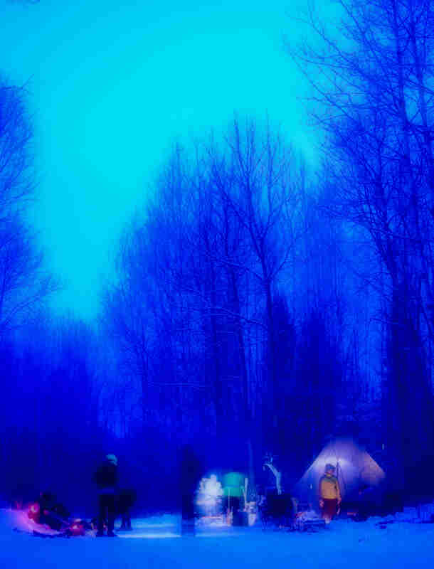 Blurry blue saturation view of people standing around at a campsite in the snow with a teepee, in the woods