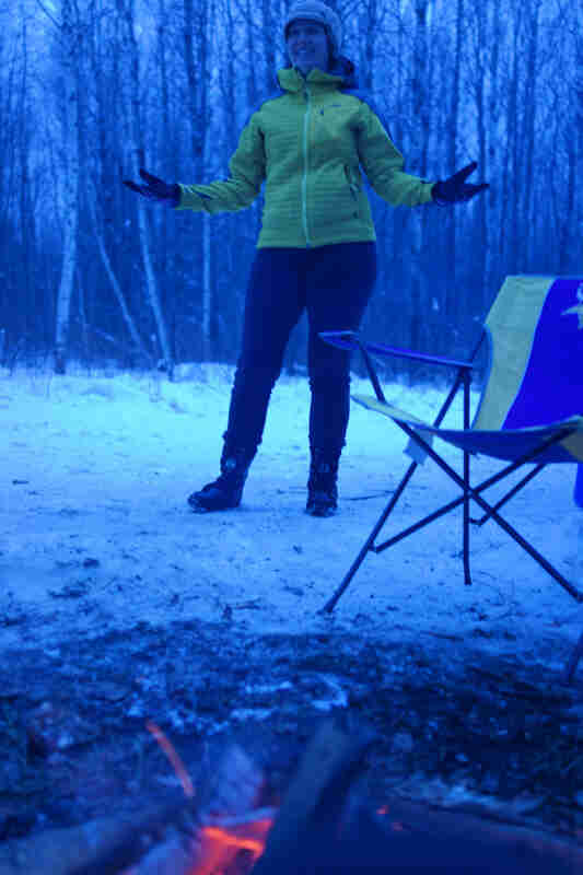 Front view of a person, dressed in winter outerwear, standing behind a campfire on the snow, in the woods