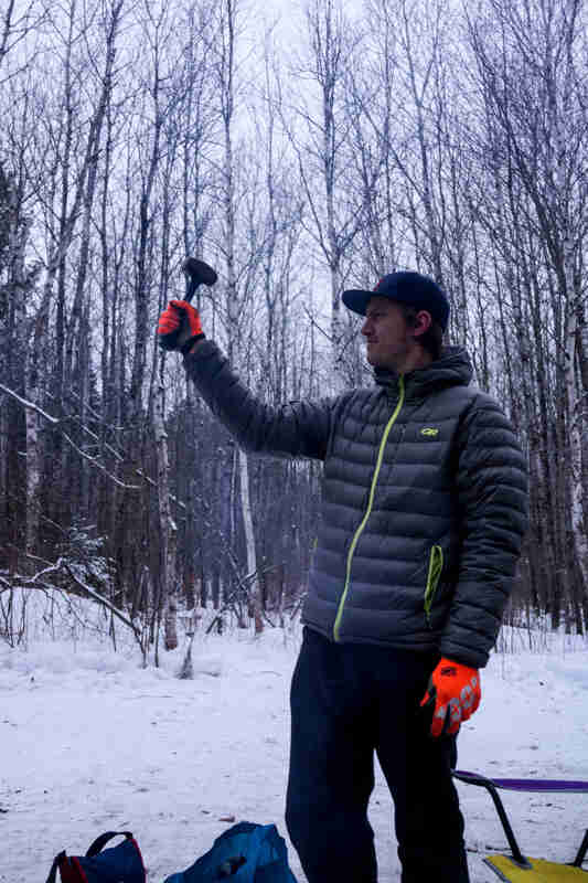A person, dressed in winter outerwear, standing and holding a hammer in their right hand, in the snowy woods