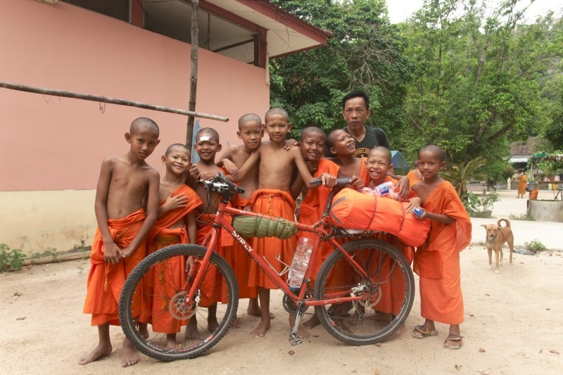 Left side view of a red Surly bike with gear, in a dirt area, with 9 children and an adult, standing behind it