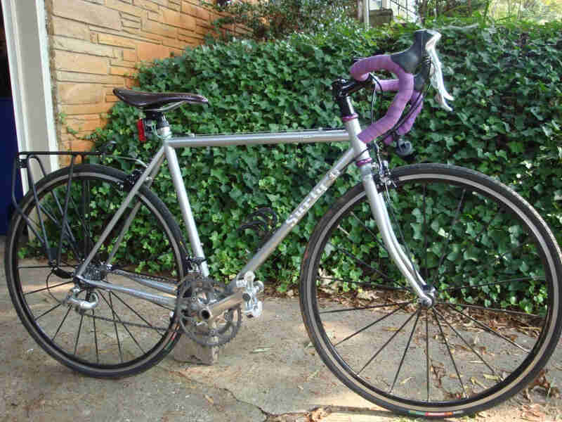 Right side view of a silver Surly bike, parked on a cement walkway, with hedge bushes behind it
