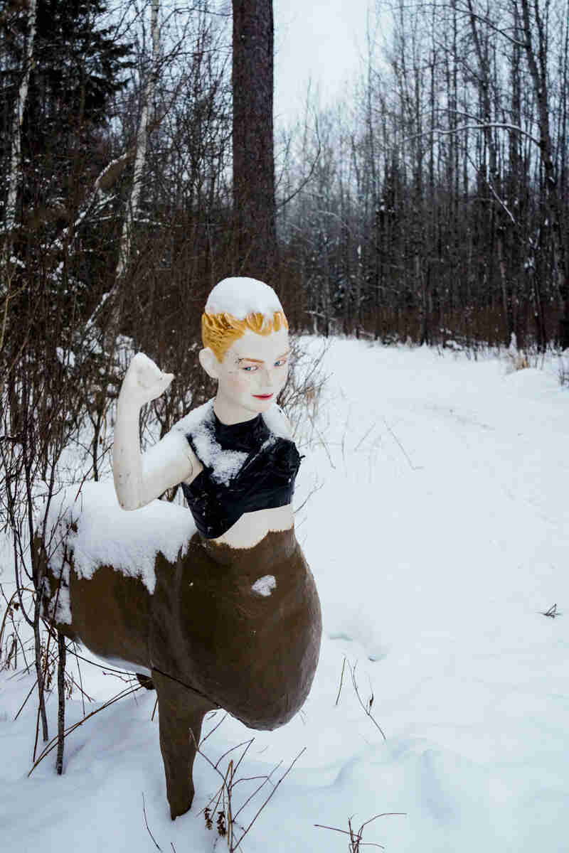 Front view of a figure with a mannequin upper half and white tail deer lower half, on a snowy trail in the woods 