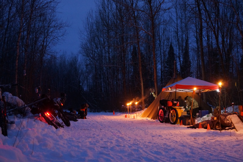 A campsite at night in the woods, with a lit up canopy, and bikes scattered around