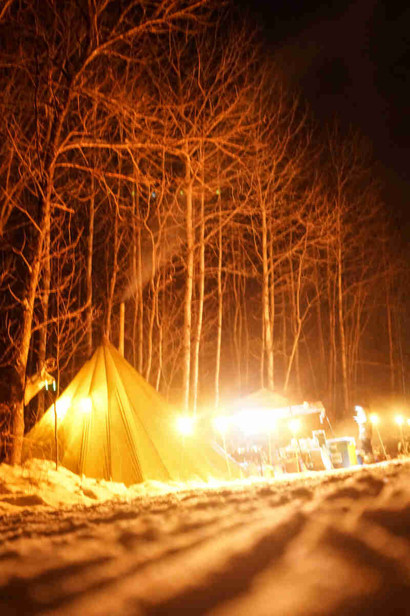 A teepee with pole lights around it and a lit up canopy, on a snowy campsite, in the woods at night