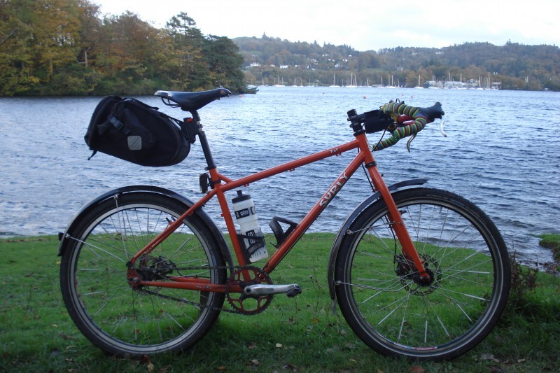 Right side view of an orange Surly Troll bike with a seat pack and fenders, parked on a flat grass bank of a lake
