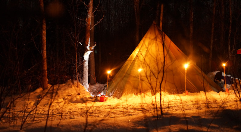 A teepee with pole lights around it, on a snowy campsite in the trees, at night