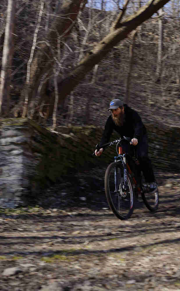 Front view of a cyclist riding down a hill on a rocky trail, along a stone wall, with trees up above in the background