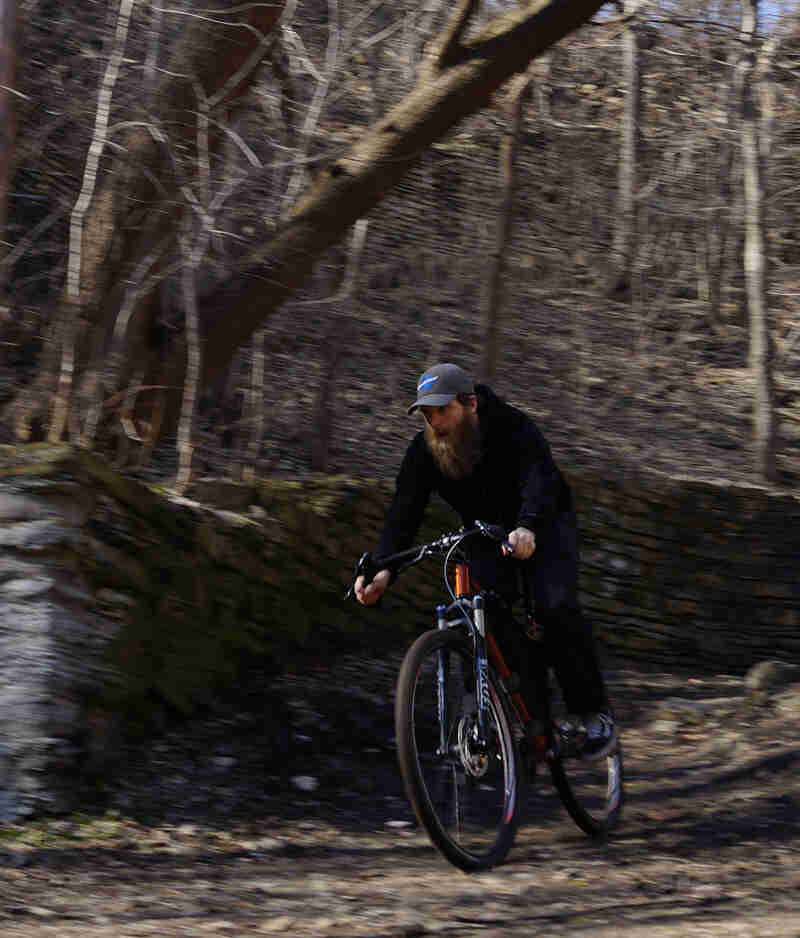 Front view of a cyclist riding down a hill on a rocky trail, along a stone wall, with trees up above in the background