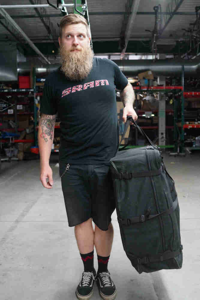 Front view of a person with a beard, standing on a warehouse floor, with a travel bag in their left hand