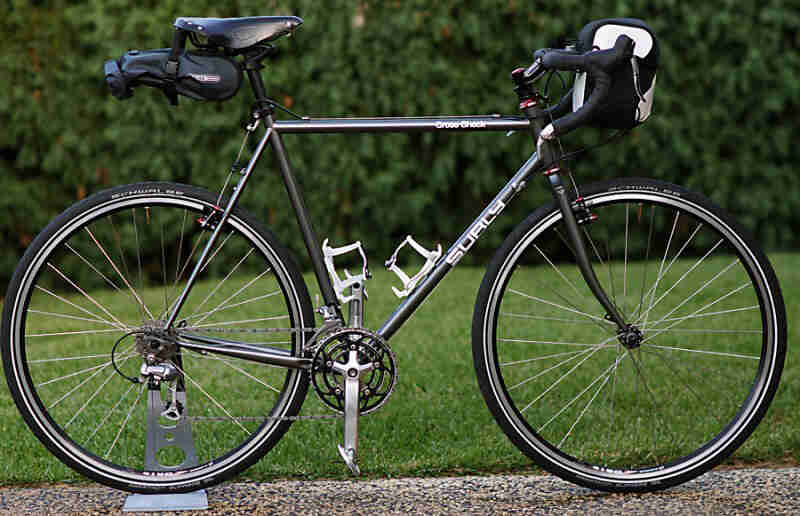 Right profile of a Surly Cross Check bike, black, on a pebble trail, with a green field and hedges in the background