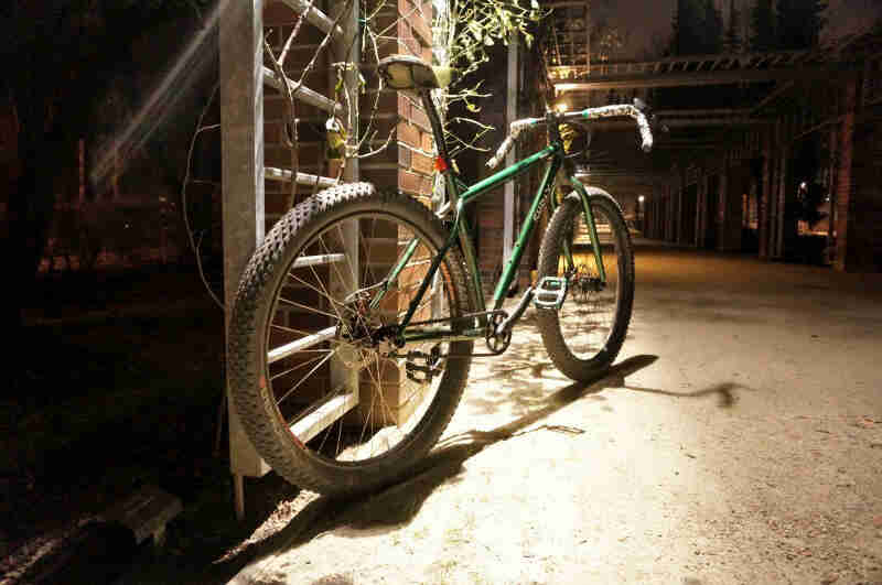 Rear view of a Surly bike, green, parked against a brick support, facing down an alley at night