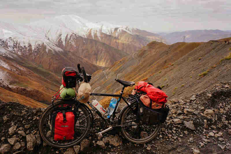 Left side view of a Surly bike, loaded with gear, parked on a rocky ledge, with snow capped mountains in the background