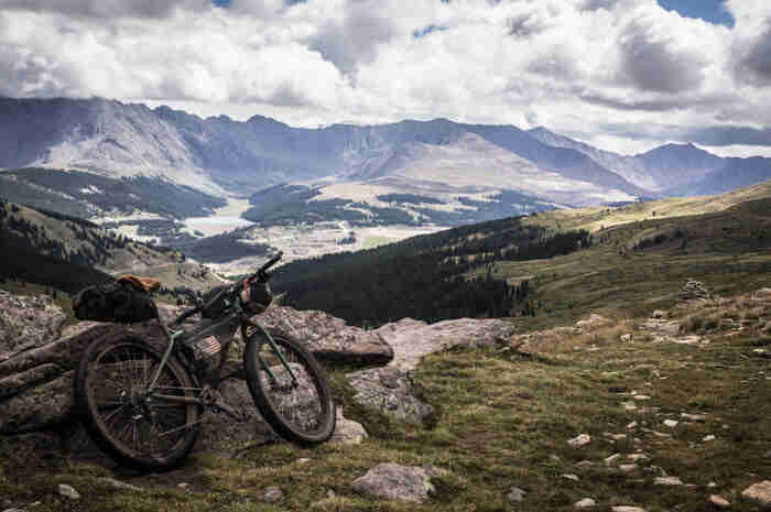 Rear, right side view of Surly Krampus bike with gear, leaning on a rock, facing grassy hills with mountains behind them