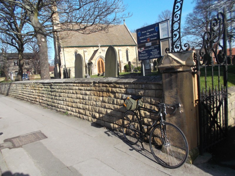 Front, right side view of a black Surly bike, parked on a sidewalk, against a stone wall with a church behind it