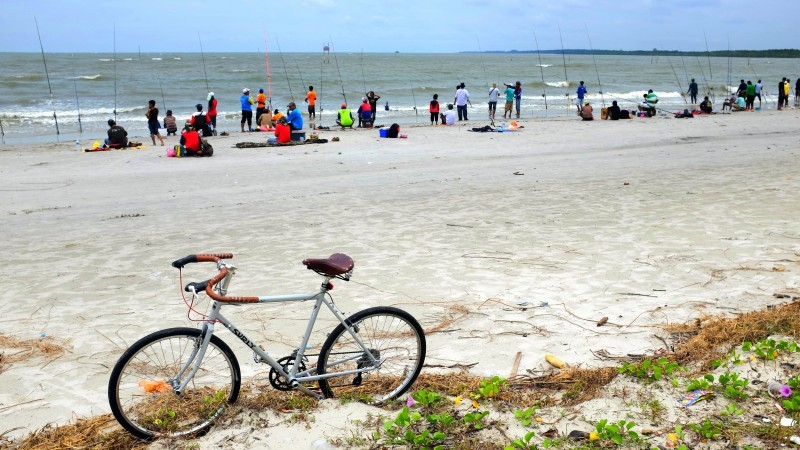 Left side view of a Surly bike, parked on the back of a sand beach, with people fishing on the shore, in the background