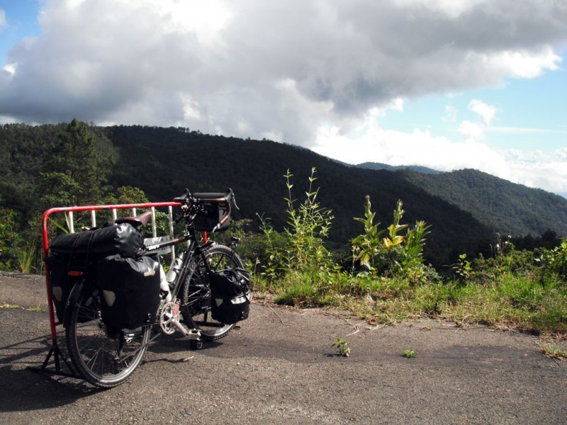 Rear, right side view of a black Surly bike loaded with gear, parked across a paved road, facing tree covered hills