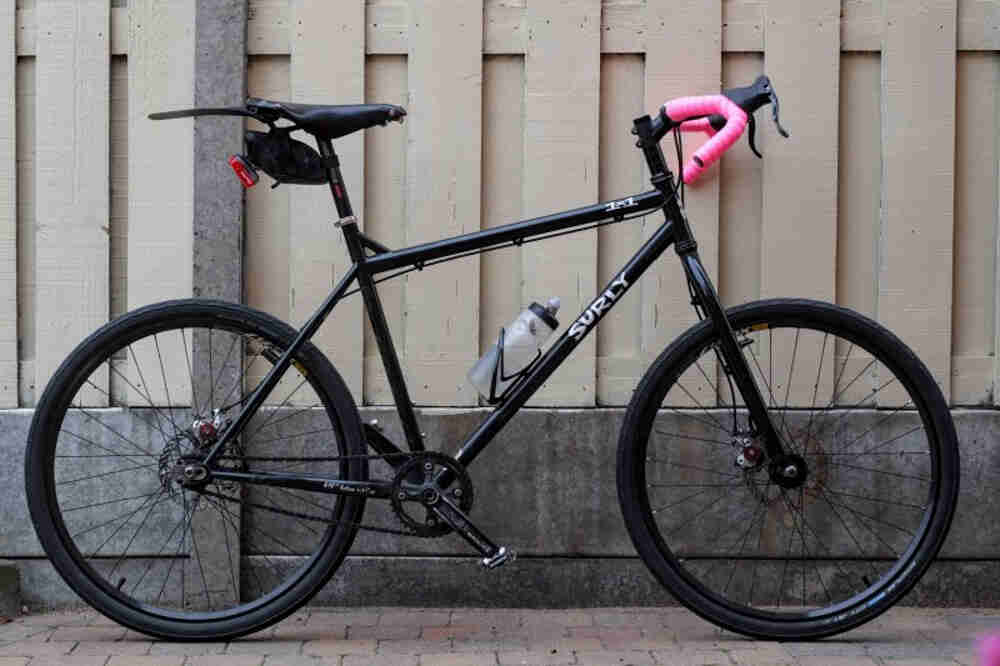 Right side view of a black Surly 1x1 bike, on a brick sidewalk next to a wood fence