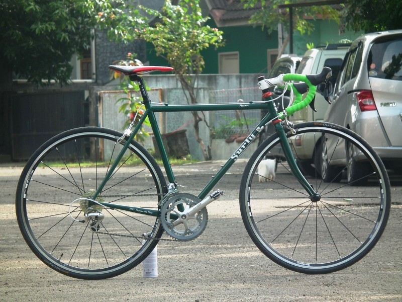 Right side view of a green Surly Pacer bike, parked in a cement lot, with cars, a wall, and a house in the background