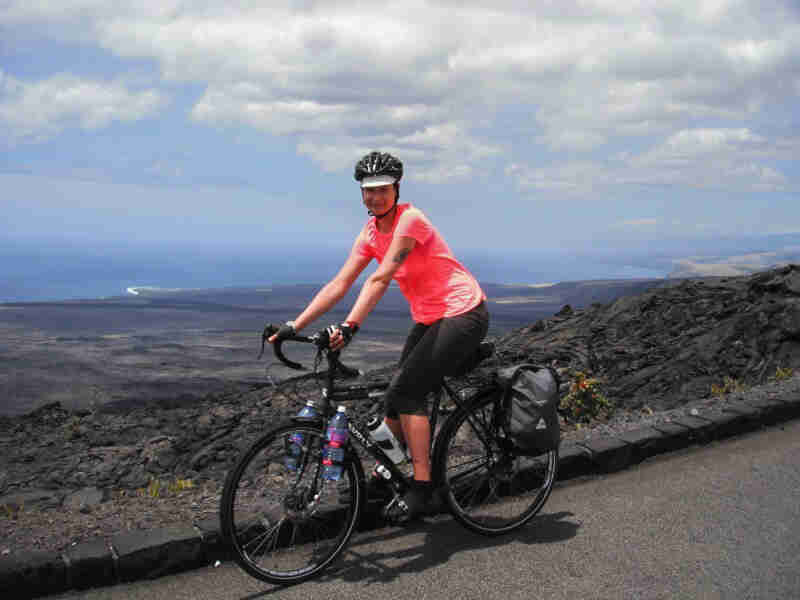 Left view of a cyclist on a black Surly bike, stopped on the side of a paved trail, with a field of volcanic rock behind